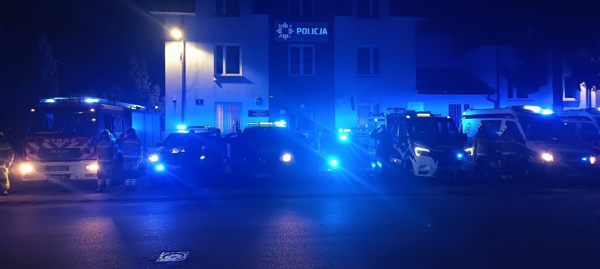 policjanci oraz funkcjonariusze innych służ stoją przy pojazdach służbowych z włączonymi sygnałami świetlnymi oddając hołd zmarłemu
