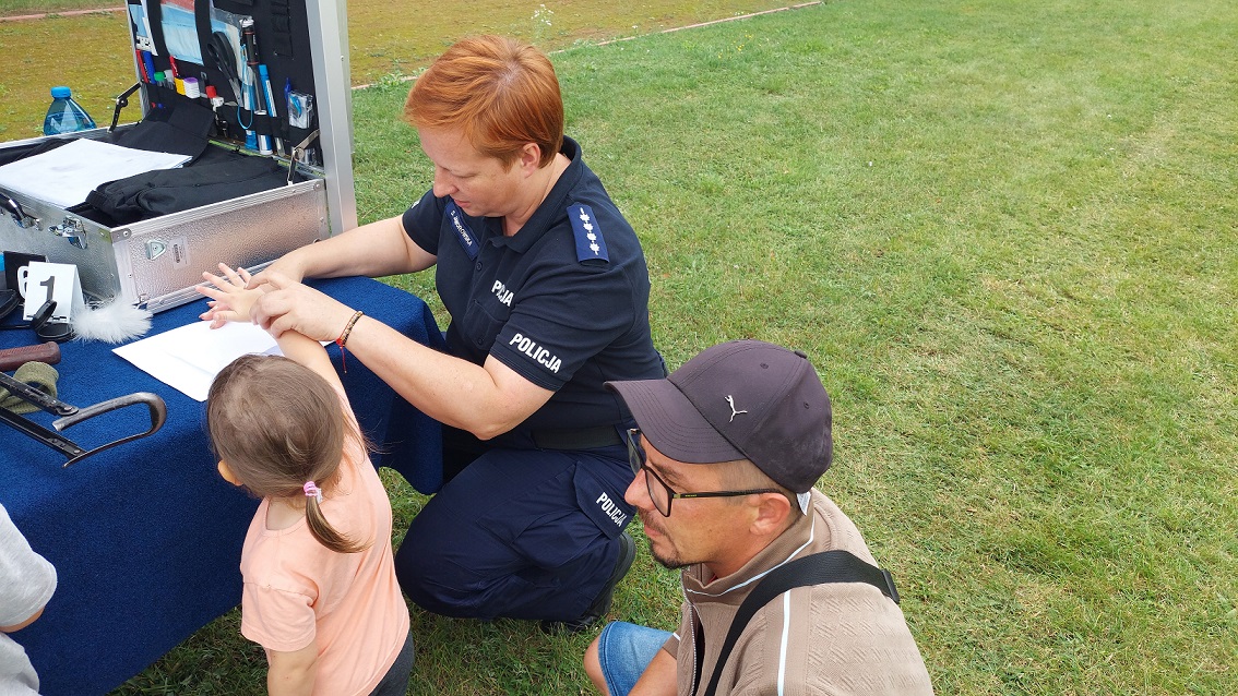 Policjantka pokazuje dziecku sposób zabezpieczenia odbitek linii papilarnych. Dzziewczynka stoi przy stole obok policjantka, która przykucnęła