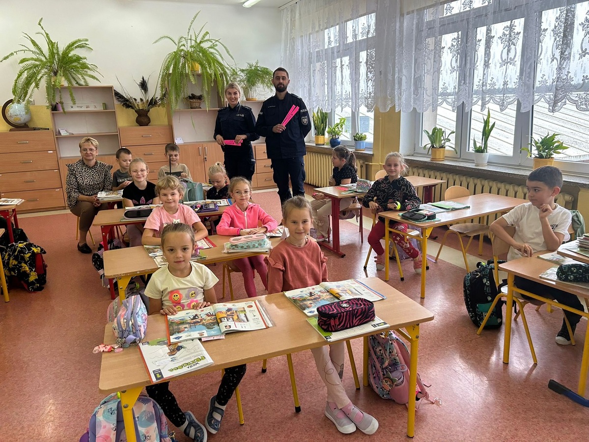 policjanci podczas spotkania z dziećmi. Uczniowie siedzą w ławkach pokazując elementy odblaskowe. Policjanci trzymaja w rękach opaski odblaskowe