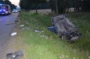 miejsce wypadku drogowego samochód na poboczu drogi leżący na dachu, za nim samochody służb ratunkowych