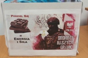 czekolady włożone do pudełka. Po lewej stronie napis Podziel się, pod spodem czekolada poniżej znak równa się energia i siła, po prawej stronie sylwetka funkcjonariusza, za nim orzeł oraz słupek graniczny. Na dole napis Bronimy Naszych Granic