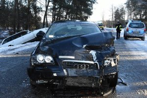 uszkodzone audi, po lewej stronie, w rowie drugi pojazd uczestniczący w zdarzeniu. Po przeciwnej stronie, w tle radiowóz