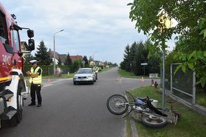 miejsce zdarzenia drogowego. Po prawej stronie motocykl. Po lewej fragment wozu strażackiego. W tle uszkodzony samochód osobowy