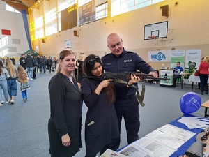 policjant wraz z dwiema kobietami prezentujący wyposażenie policji. Jedna z kobiet trzyma broń