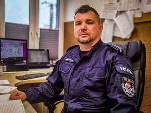 policjant siedzący za biurkiem. Za nim w tle monitory