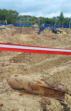 niewybuch leżący na ziemi. Przed nim taśma zabezpieczająca. W tle koparka