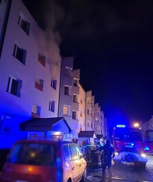 miejsce pożaru. Dym wydobywający się z okna, pojazdy służb ratunkowych pod blokiem oraz ratownicy