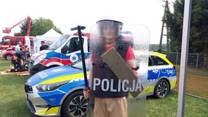 dziecko ubrane w umundurowanie PZ. Za nim widoczny radiowóz i karetka pogotowia