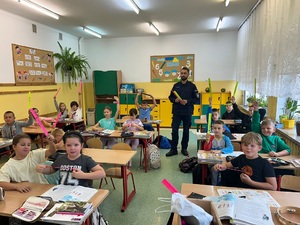 policjant na spotkaniu z dziećmi. Dzieci siedzą w ławkach trzymając w rękach opaski odblaskowe