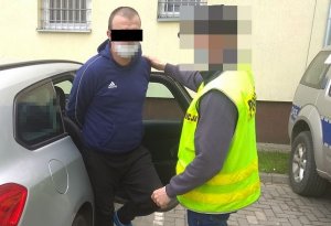 policjant doprowadza zatrzymanego 38-latka. Mężczyzna wysiada z samochodu